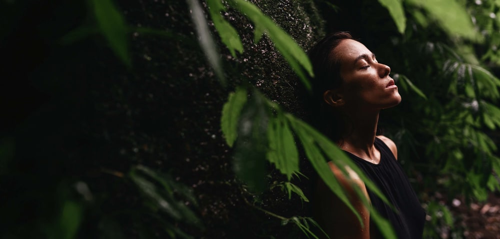 A woman with eyes closed leaning against a mossy rock wall among green tropical leaves