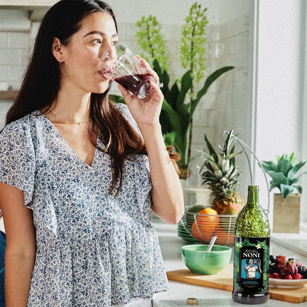A woman drinking a glass of Tahitian Noni juice in a bright kitchen, with the bottle on the counter