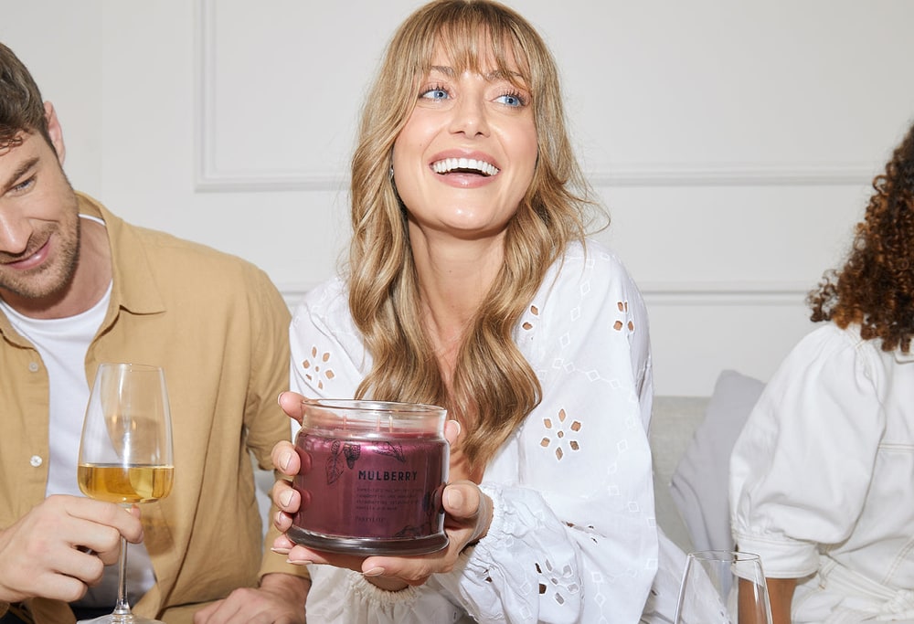 A smiling woman holding a PartyLite jar candle while friends with wine glasses sit beside her on a sofa