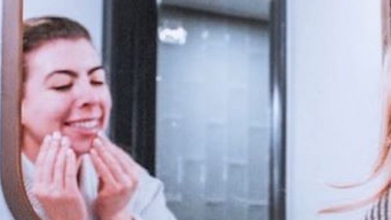 A smiling woman in a bathrobe massaging cleanser into her face, reflected in a bathroom mirror