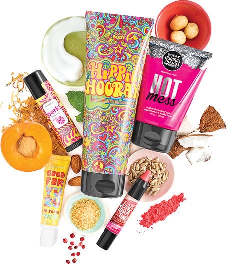 A spread of colorful Perfectly Posh body crème, hand crème, lip scrub and skin stick surrounded by apricot, coconut, almonds and other ingredients