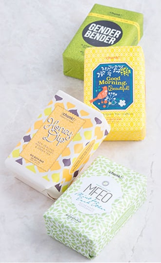Four wrapped Perfectly Posh Chunk bath bars in green, yellow and white patterned papers on a marble surface