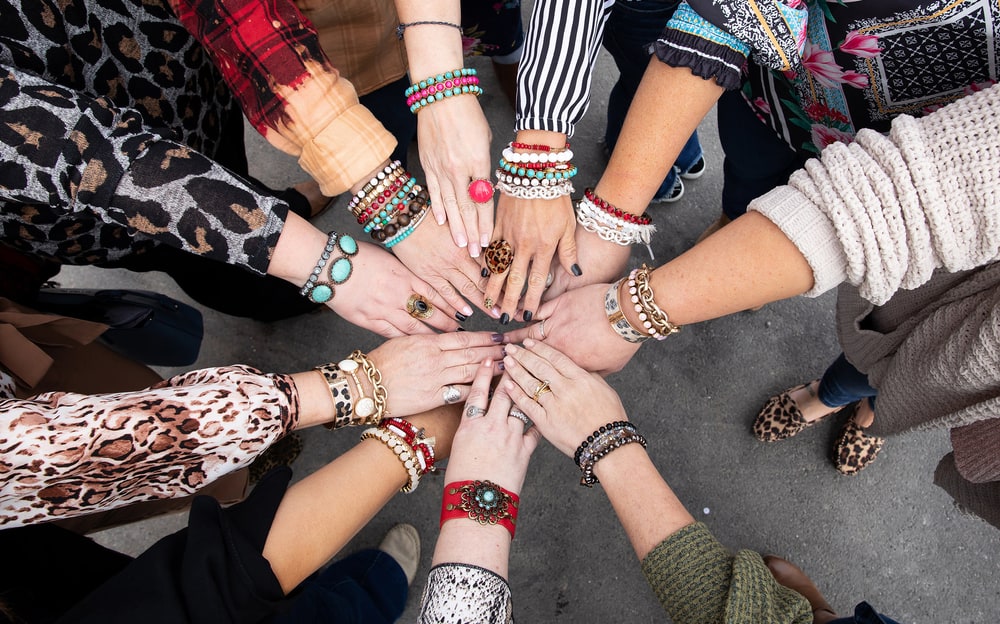 A circle of women's hands stacked together, wrists covered in beaded, leather and metal Plunder bracelets and rings