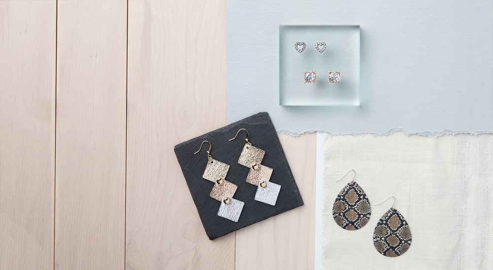 A flat lay of Plunder earrings: hammered metal squares on slate, snakeskin-print teardrops on linen and small studs on a clear block