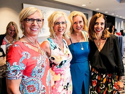 Four smiling women in bright dresses wearing pendant necklaces at an indoor event