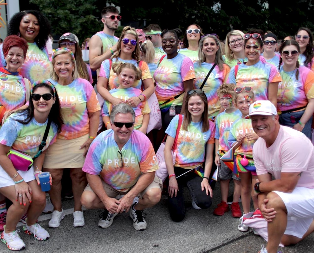 A large group of Pure Romance staff and families in matching tie-dye Pride T-shirts posing together at a street parade