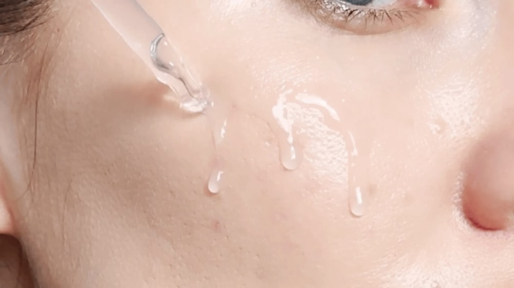 Clear hydrating serum dripping from a glass dropper onto a woman's cheek