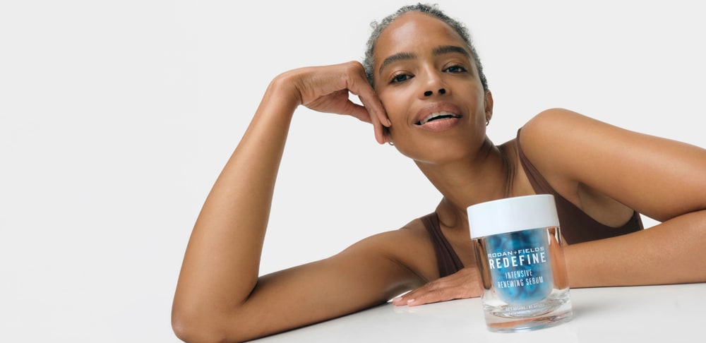 Smiling woman with grey hair resting her arms on a white table beside a jar of Redefine Intensive Renewing Serum capsules