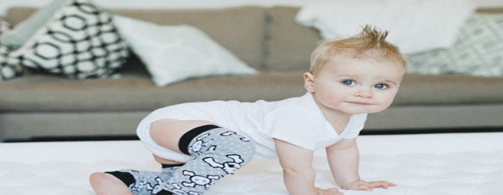 A toddler in a white bodysuit and printed leg warmers crawls across a white bed in front of a sofa
