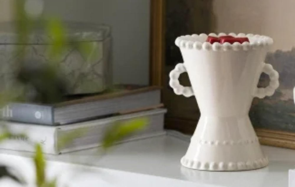 A white beaded Scentsy warmer with melted red wax in its dish, sitting on a shelf beside stacked books and a framed landscape