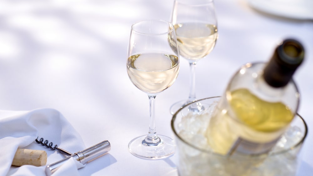 Two glasses of white wine on a white tablecloth beside a bottle in an ice bucket and a corkscrew