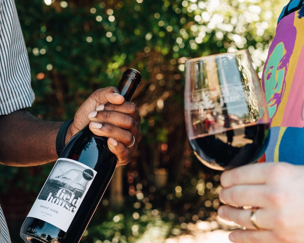 A Consultant holds a bottle of Fieldhouse Pinot Noir while a guest raises a glass of red wine outdoors