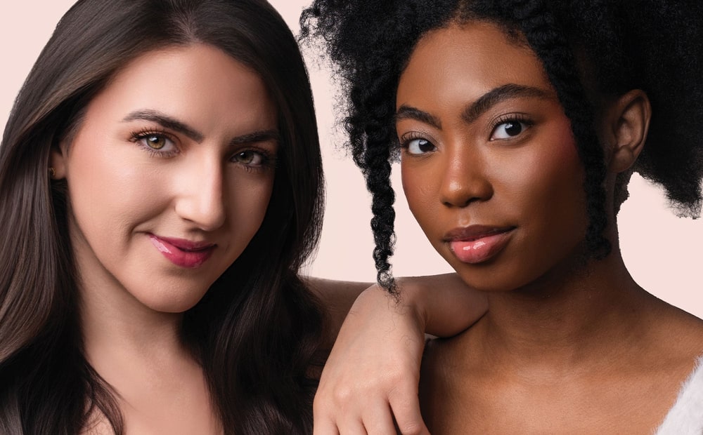 Two smiling women with glossy pink lips posing together against a soft pink background