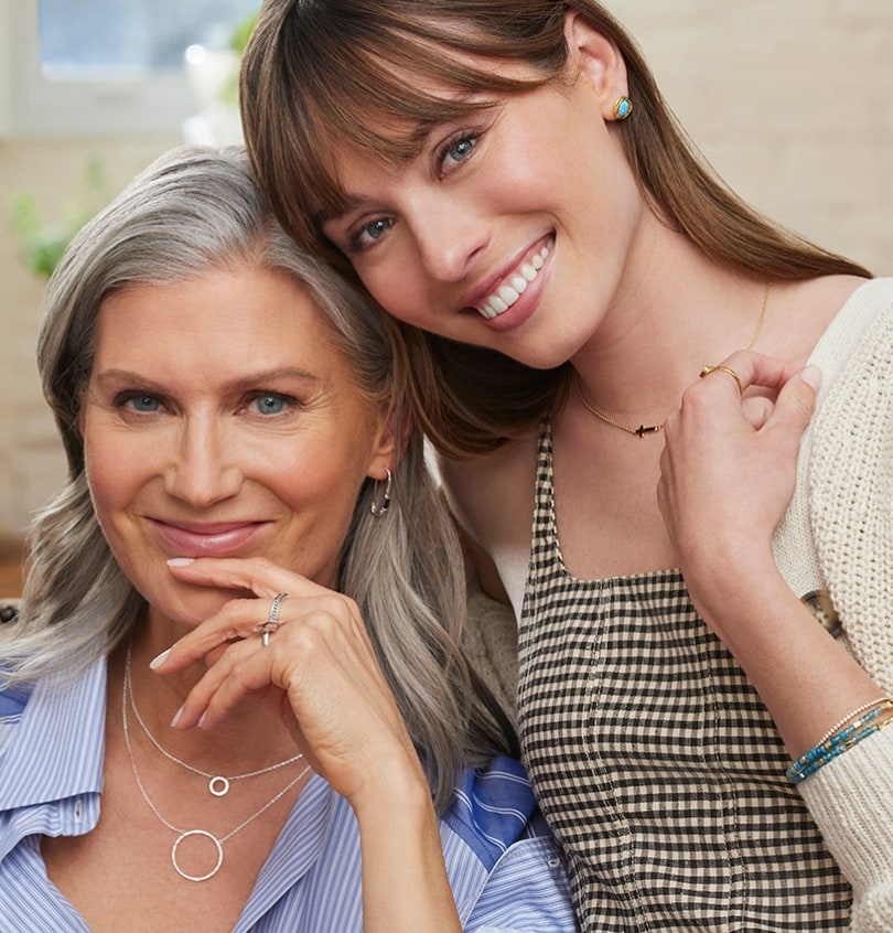 An older and a younger woman hugging, wearing delicate silver and gold necklaces and bracelets