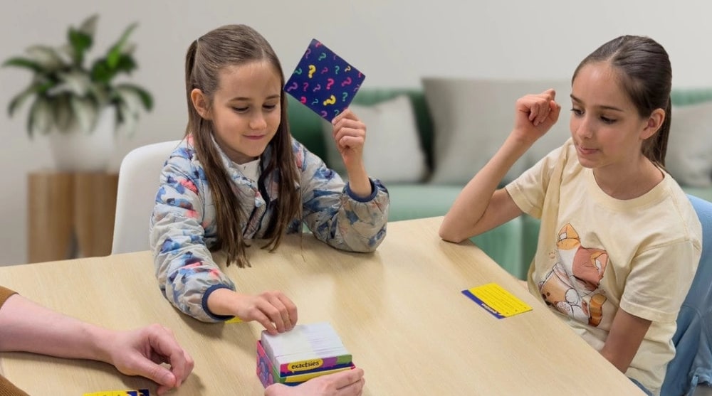 Two girls play a card guessing game at a family table, one holding up a card