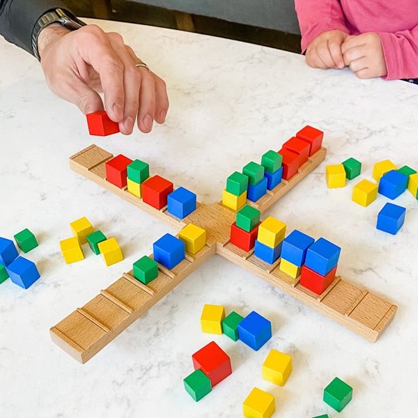 An adult and a child stack colored wooden cubes on a cross-shaped wooden balance beam