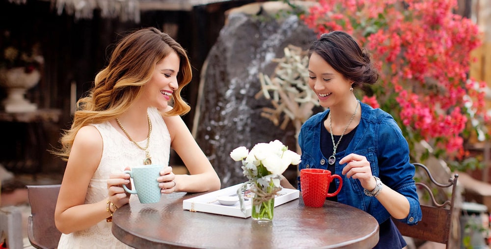 Two women laugh over coffee at an outdoor cafe table, both wearing pendant necklaces