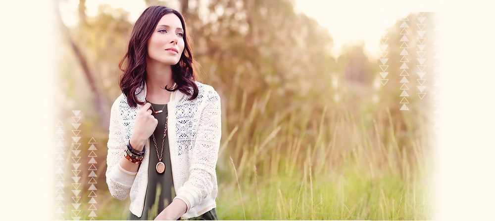 A woman in a cream lace cardigan wears a long locket necklace and stacked bracelets in a sunlit field