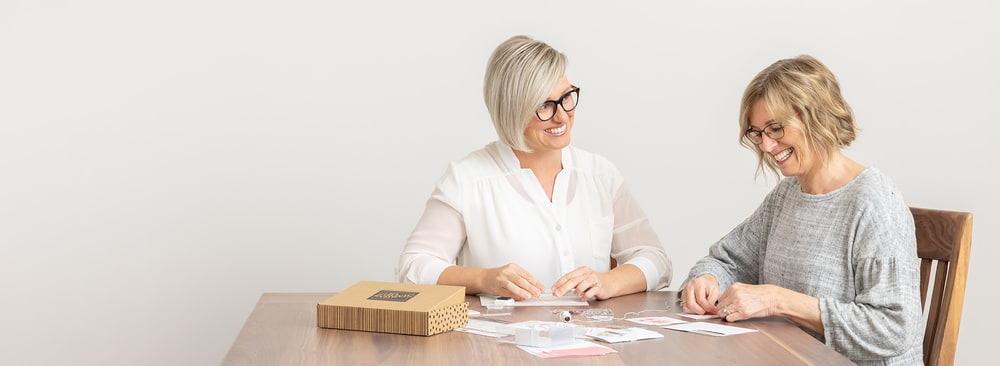 Two women assembling handmade cards at a wooden table next to a Paper Pumpkin kit box