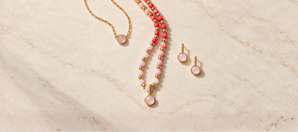 A gold chain necklace with a rose quartz bezel, a pink beaded pendant necklace and matching bar stud earrings laid on cream marble