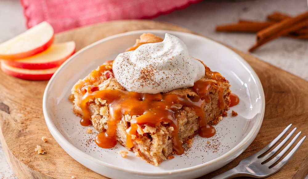 A square of apple cake topped with whipped cream and caramel sauce on a white plate, with apple slices and cinnamon sticks