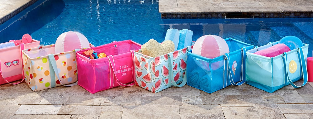 A row of brightly printed Thirty-One utility totes filled with beach balls, towels and sun hats beside a swimming pool