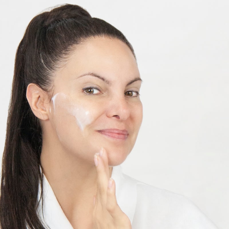 A smiling woman with her hair pulled back applying a white moisturizer to her cheek