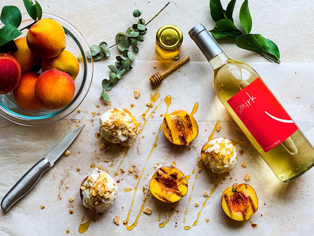 A bottle of Smirk Muscat lying beside grilled peaches topped with cream, honey and a bowl of fresh peaches