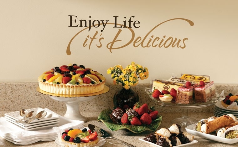 A dessert table with a fruit tart, cakes and strawberries beneath vinyl wall lettering reading Enjoy Life, it's Delicious
