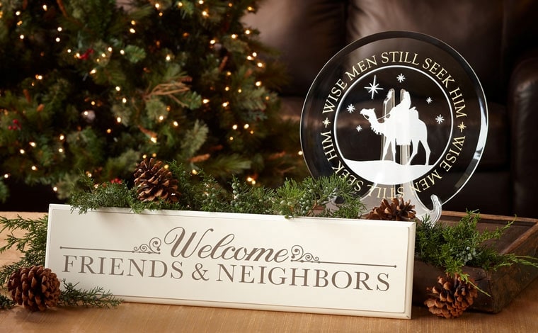 A white wooden plaque lettered Welcome Friends and Neighbors beside a glass ornament with a nativity design, among pine branches