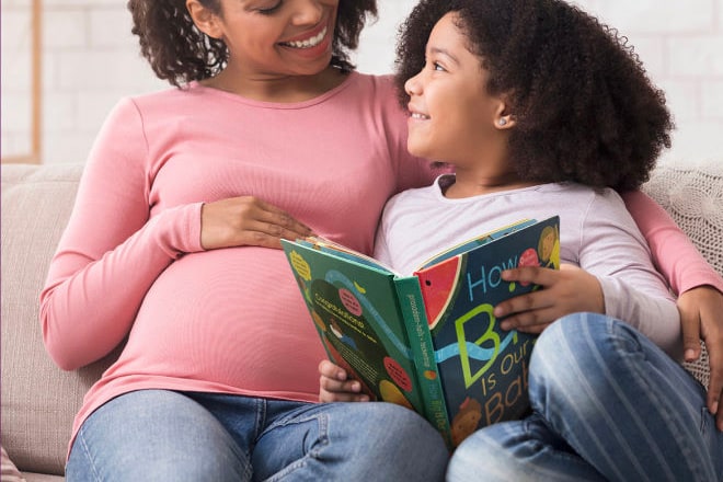 A smiling mother on a sofa with her arm around her daughter, who holds open a brightly illustrated picture book