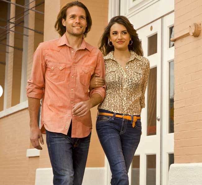 A man in an orange shirt and a woman in a leopard print blouse walking arm in arm in dark jeans
