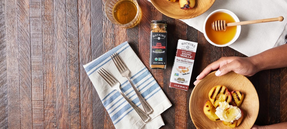 Grilled peaches with cream and honey on a wooden table beside Watkins Organic Ground Cinnamon and Baking Vanilla