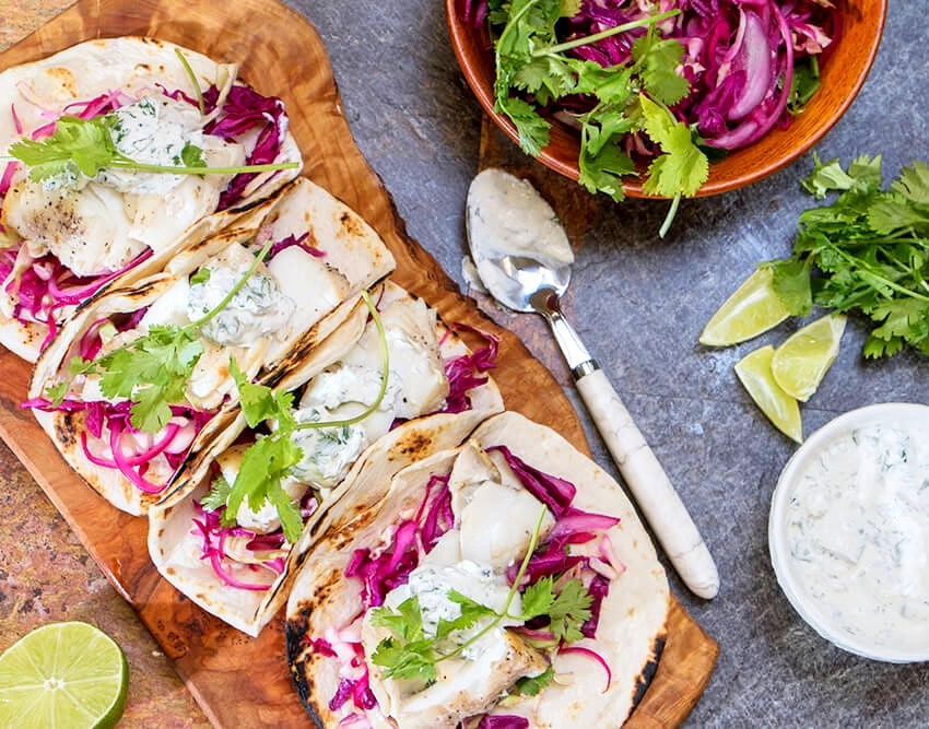 Grilled fish tacos with red cabbage slaw, cilantro and a creamy herb sauce on a wooden board, with lime wedges beside them