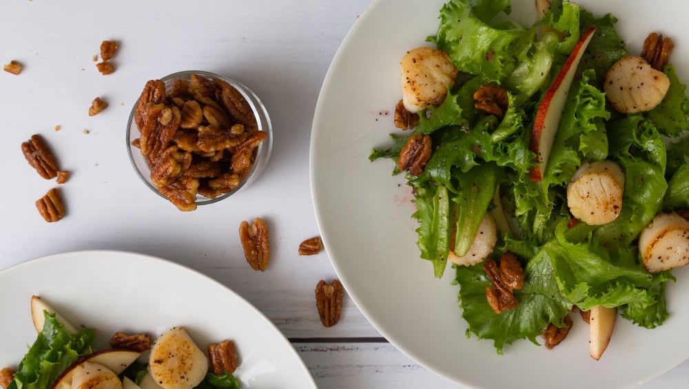 A green salad with seared scallops, sliced pear and candied pecans on white plates, with a small glass of pecans