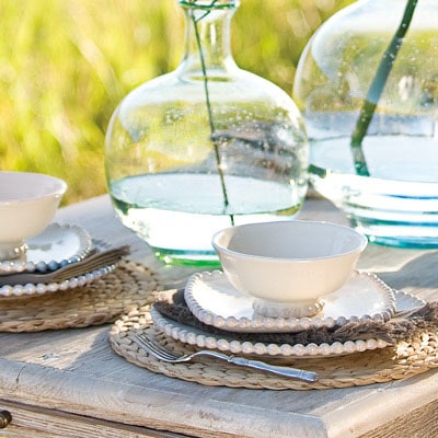 Cream beaded-rim plates and bowls on woven chargers, with large green glass bottles on a rustic outdoor table