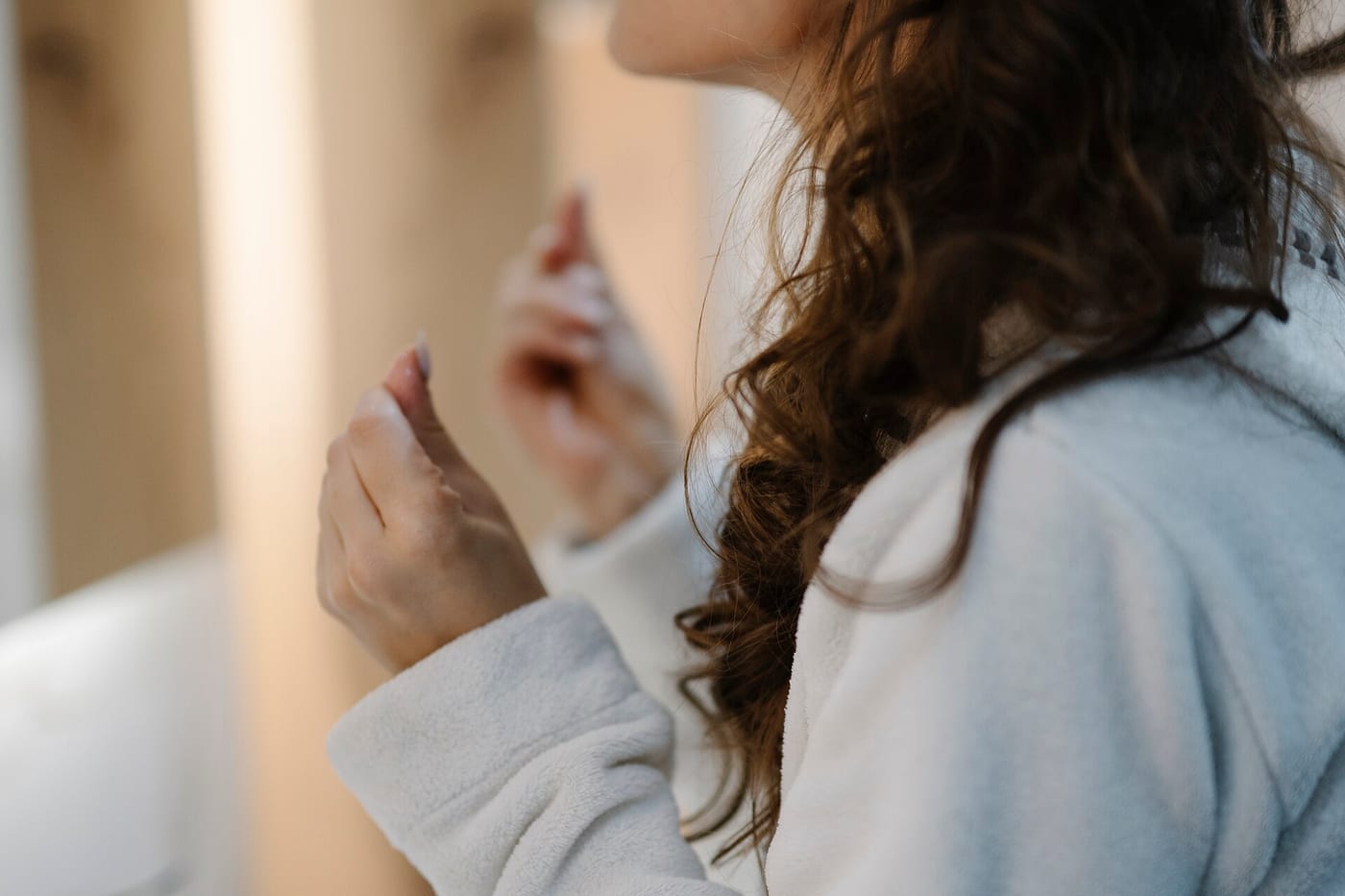 A woman in a white robe applies a skin care product in front of a bathroom mirror